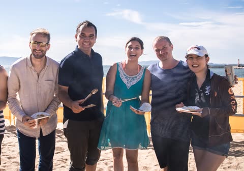 A group of five people holding paper plates and eating utensils smile while standing together on a beach.
