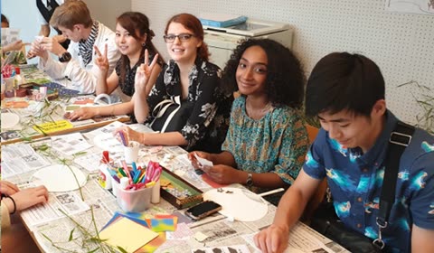 A group of young adults sits around a long table covered with newspapers and craft supplies during an indoor activity.
