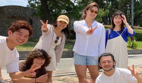 A group of six people smile and make peace signs with their hands while posing outdoors in front of a stone wall.