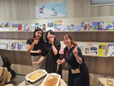 Three women smile and pose with chopsticks and noodles in front of a wall of travel brochures.