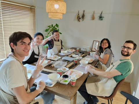 Five people wearing aprons and gloves smile while sitting at a wooden table preparing food in a sunlit room.