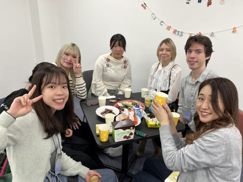 Six people pose for a group photo, sitting around a black table with snacks in a room decorated with a Christmas banner.