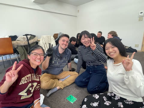 Four people sit on the carpeted floor of a room, smiling and making peace signs at the camera.