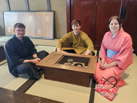 Two men and a woman in kimonos sit on a tatami mat floor around a wooden table with a built-in hearth.